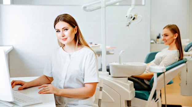 dental assistant working at computer with patient in exam chair in the background
